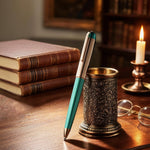 Teal pen with decorative holder on a wooden desk with books and candle in the background